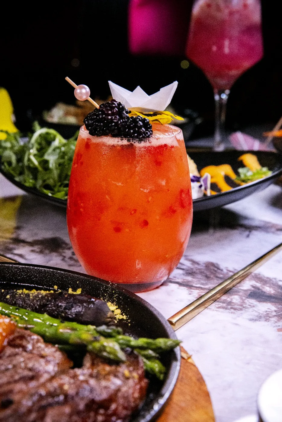 An orange Mousai cocktail with garnish beside dishes at the restaurant